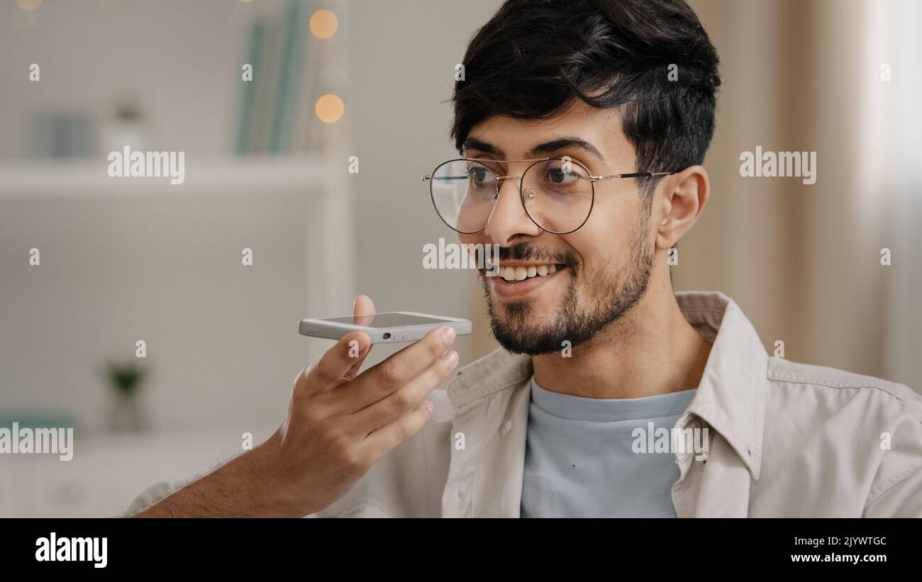 Freundlicher junger arabischer Mann, der die Spracherkennungsnachricht auf einem zu Hause sitzenden Lautsprechertelefon aufzeichnete. Bärtiger Kerl mit Brille mit virtuellen Assistenten App-Sets Stockfoto
