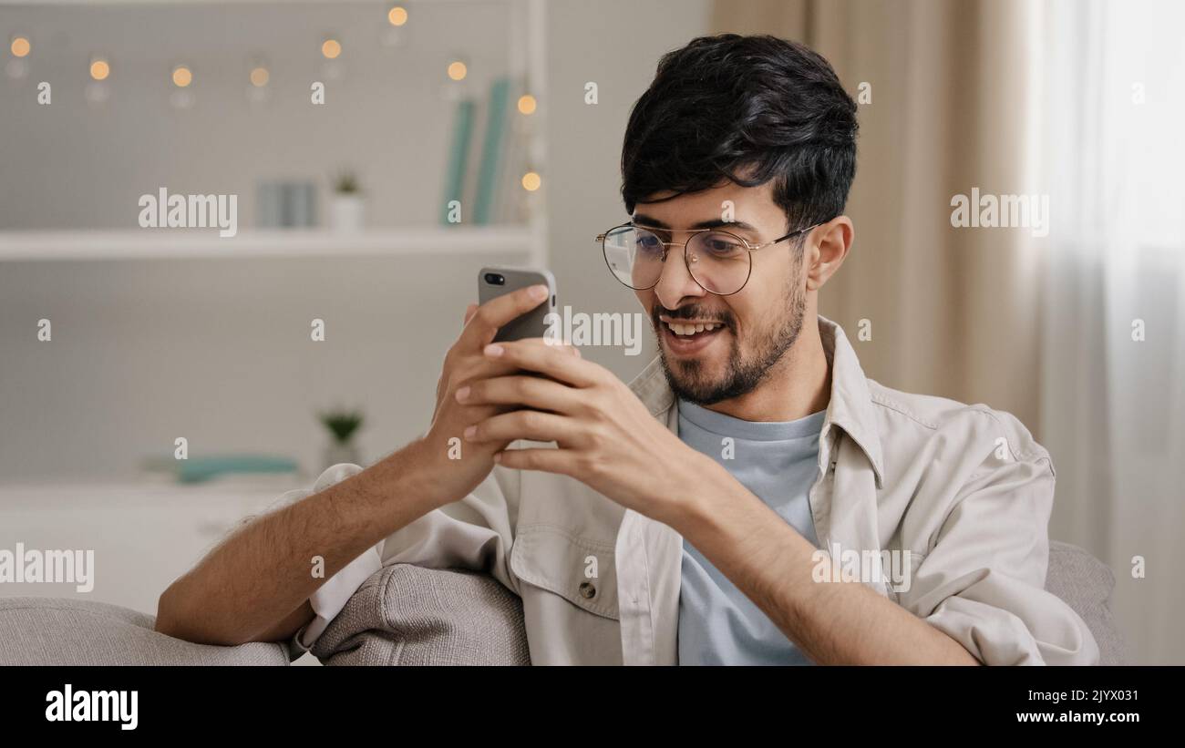 Junger 30s araber bärtiger Mann Millennial Kerl in Brille sitzt zu Hause auf der Couch Blick auf Handy macht ja Geste immer Angebot gewinnen gute Nachrichten Stockfoto
