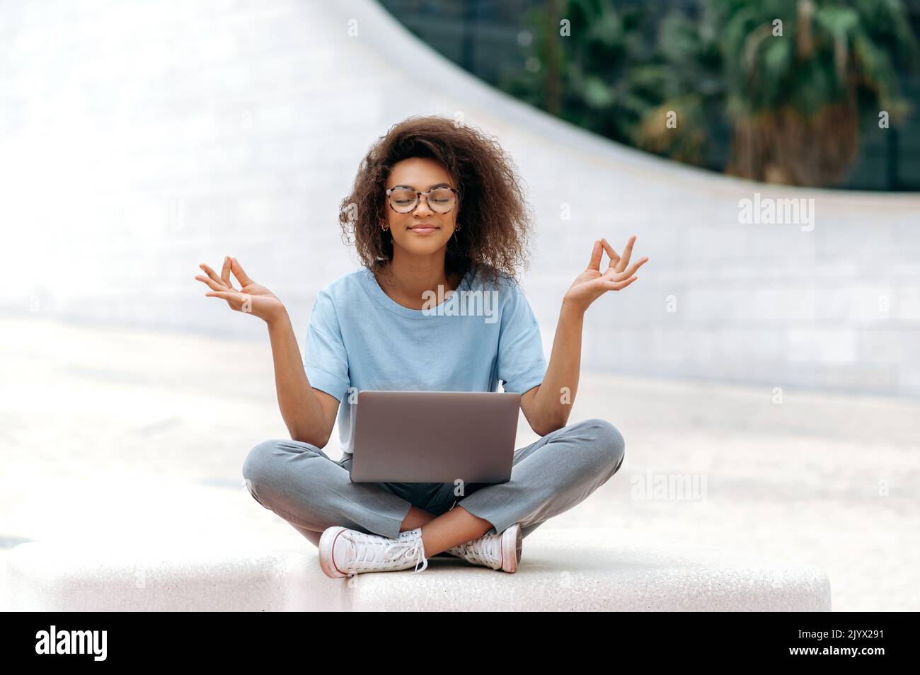 Friedliche Ruhe entspannte afroamerikanische junge Frau mit Brille, Büroangestellte, sitzt in einer Lotusposition im Freien in der Nähe des Geschäftszentrums, meditiert, entspannt, schließt ihre Augen.Pause bei der Online-Arbeit Stockfoto