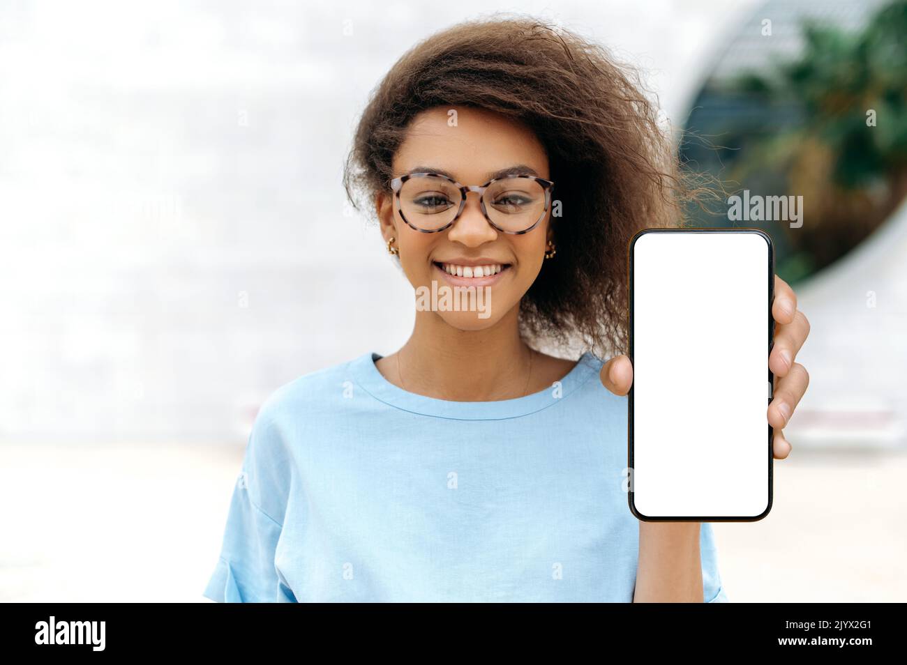 Trendy positive Lovely mixed race junge lockige haarige Frau mit Brille, steht im Freien, zeigt Smartphone mit leerem weißen Mock-up-Bildschirm, schaut auf die Kamera, lächelnd glücklich. Modellkonzept Stockfoto