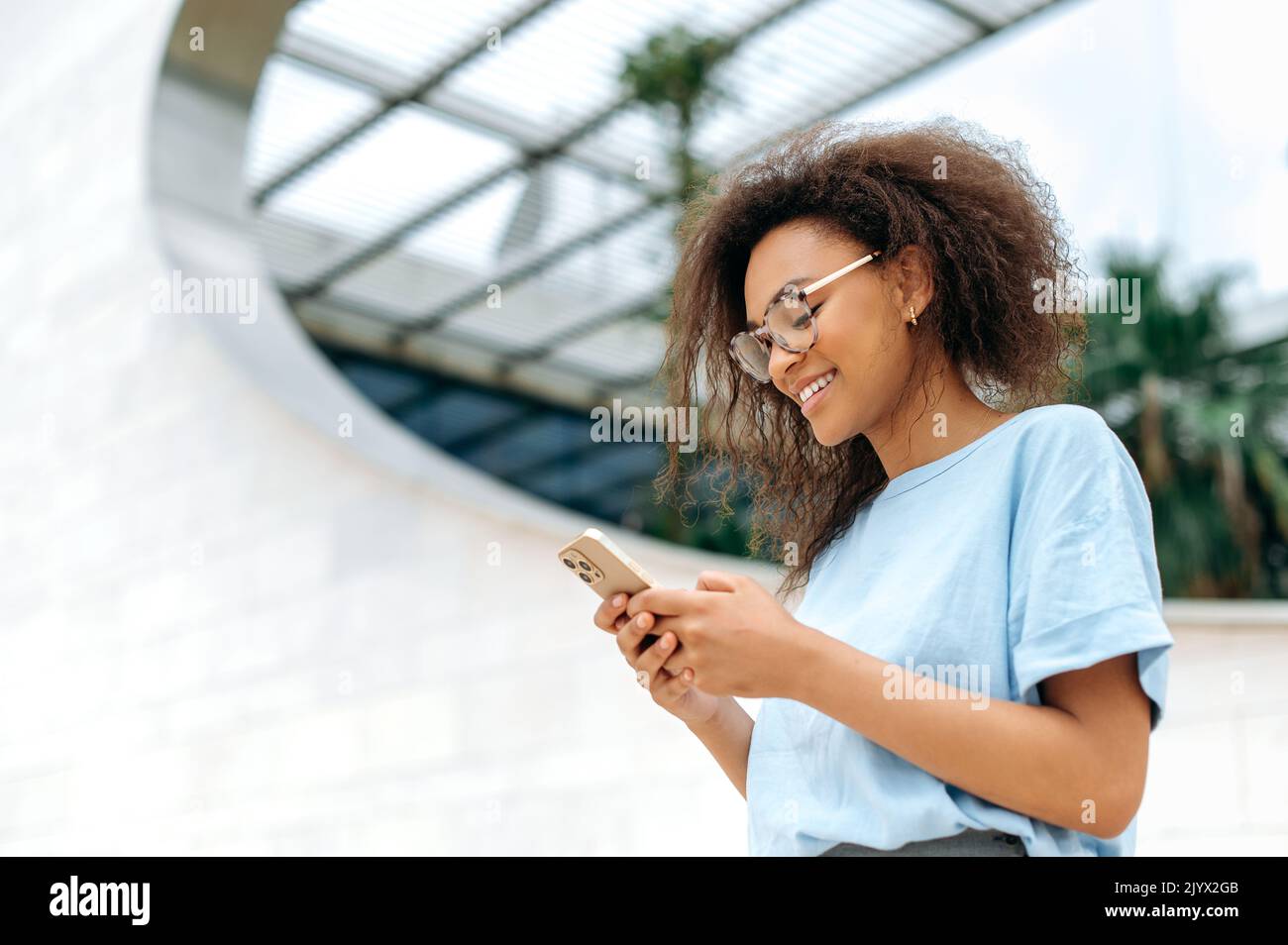 Online-Chat. Positive afroamerikanische junge Frau mit lockigen Haaren, stilvoll gekleidet, mit ihrem Smartphone im Freien stehend, Messaging mit Freunden in sozialen Netzwerken, beantwortet E-Mails und lächelt Stockfoto
