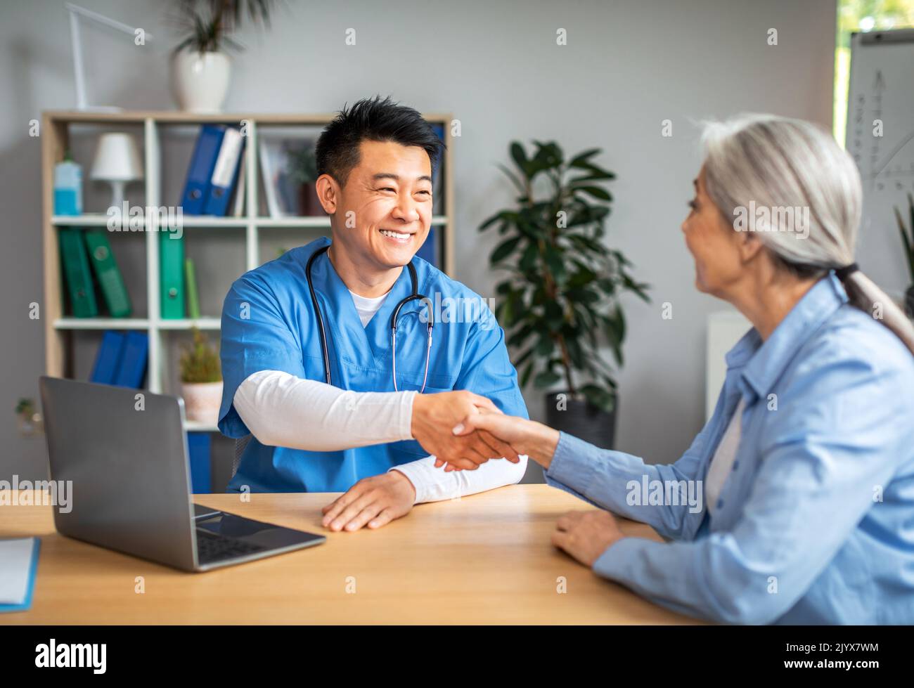 Lächelnder reifer chinesischer Mann Arzt schüttelt sich die Hände mit einer älteren kaukasischen Frau in der Klinik Stockfoto