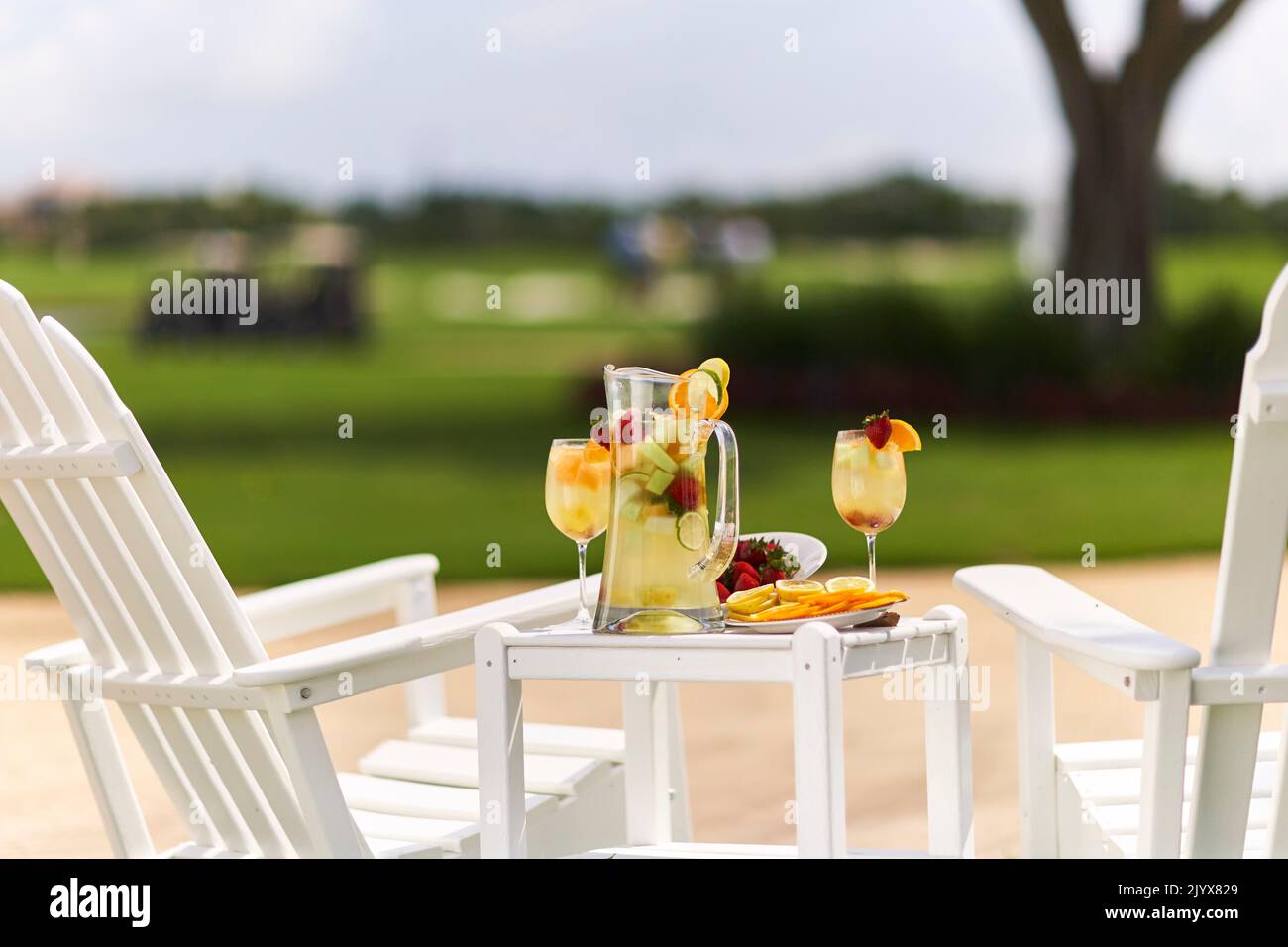 Limonade im Glaskrug mit Orangen in Scheiben und Limetten. Garniert mit Erdbeeren. Draußen auf einem kleinen Tisch mit zwei weißen Adirondack-Stühlen. Stockfoto