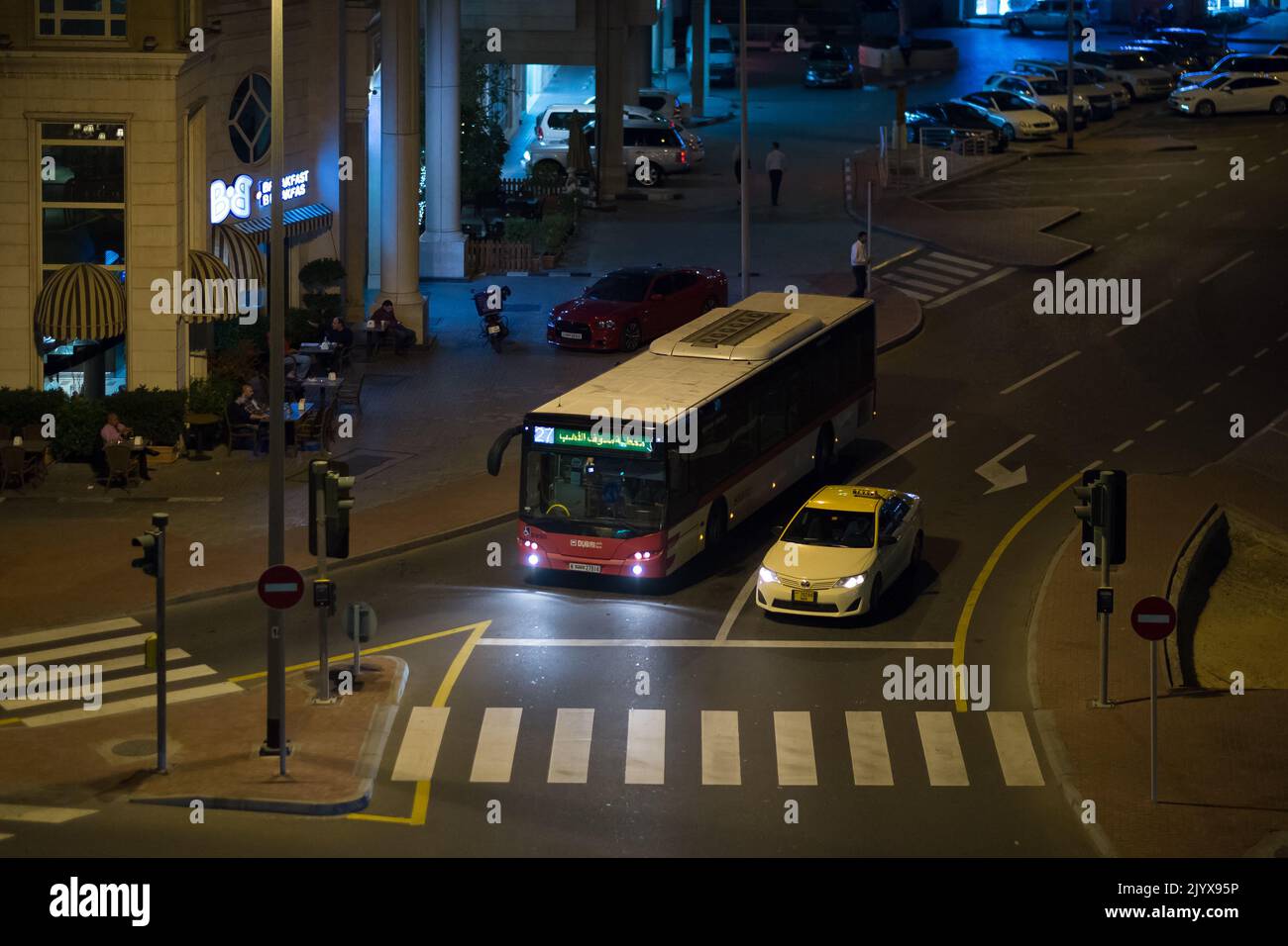 Dubai Bus und Taxi halten an einer Ampel an einem späten Abend in der Innenstadt. Der Fußgängerüberweg und die Insel sind leer. Nur wenige Leute liegen an Tischen und Stühlen vor einem Café. Stockfoto