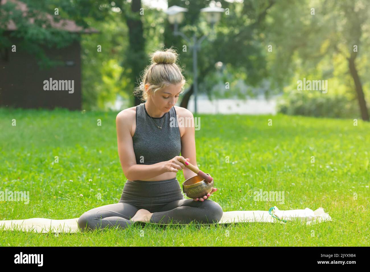 Eine schöne Frau sitzt auf dem grünen Gras, hält eine Meditationsschale, treibt einen Stock darauf. Stockfoto