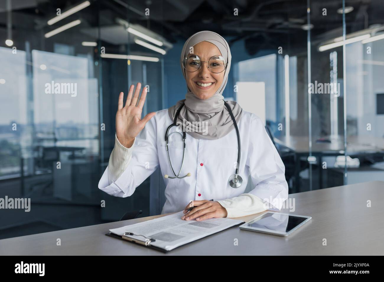 Online-Konsultation mit Videoanrufen, Frau in Hijab, muslimischer Arzt, der die Kamera anschaut und mit winkender Grußgeste winkt, Ärztin, die Patienten aus der Ferne konsultiert und in einem modernen Klinikbüro arbeitet Stockfoto