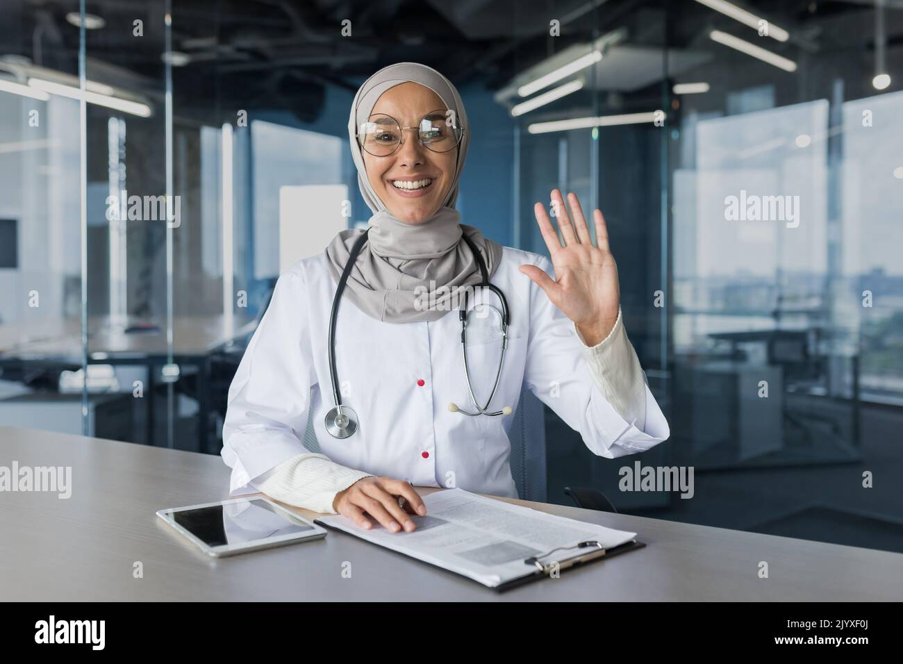 Online-Konsultation mit Videoanrufen, Frau in Hijab, muslimischer Arzt, der die Kamera anschaut und mit winkender Grußgeste winkt, Ärztin, die Patienten aus der Ferne konsultiert und in einem modernen Klinikbüro arbeitet Stockfoto