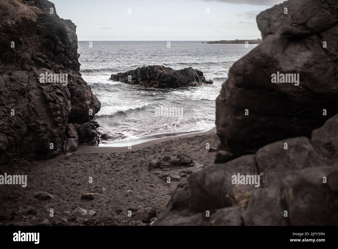 Schwarzer Sandstrand, versteckt zwischen verschwommenen vulkanischen Felsen. Dramatische Landschaft Stockfoto