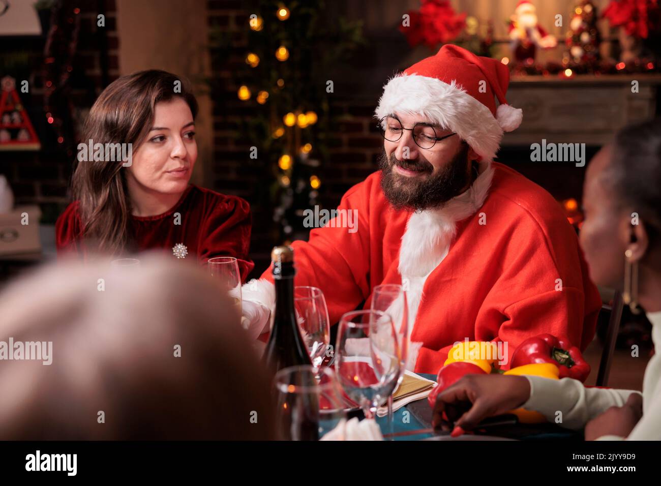 Ehemann im weihnachtsmann-Kostüm feiert weihnachten mit Frau und Eltern und trinkt Sekt am festlichen Esstisch. Winterfeiertag, Familientreffen Stockfoto