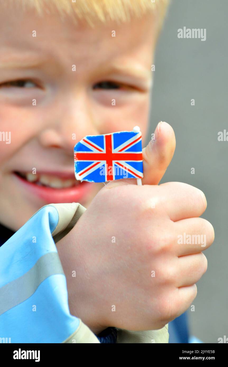 Der blonde Bursche hält die Union Jack-Flagge aus Papier, sein lächelndes Gesicht ist im Hintergrund unscharf. Stockfoto