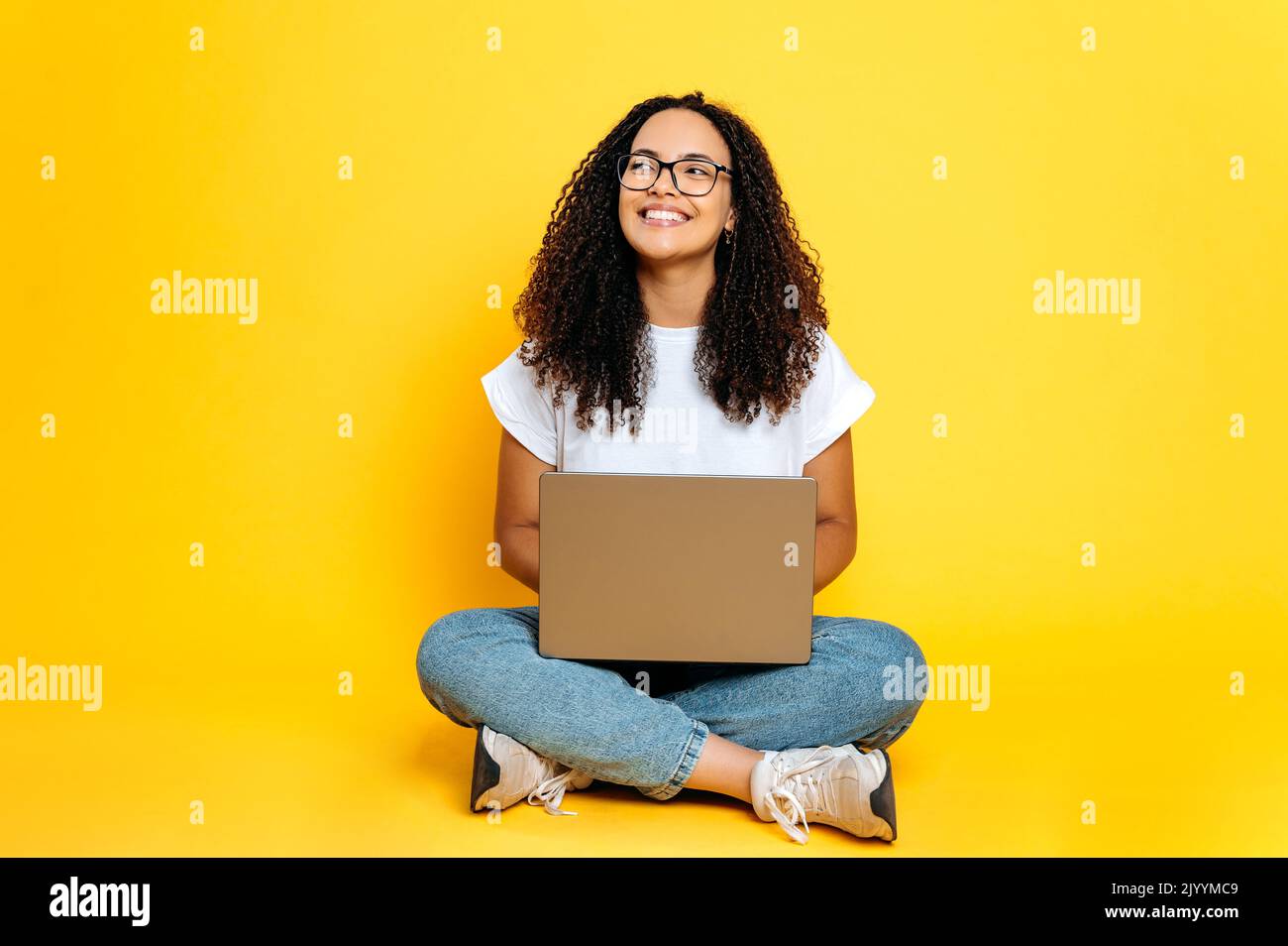 In voller Länge Foto von positiven fröhlich kreative junge brasilianische oder hispanische Frau mit Brille, Freiberufler oder Designer, sitzen auf isolierten orangefarbenen Hintergrund mit Laptop, sieht zur Seite, träumen, lächelt Stockfoto