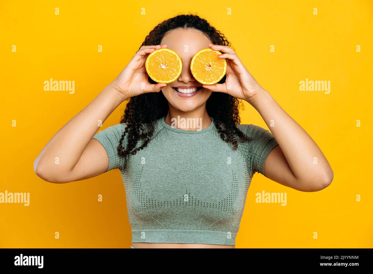 Gesunde Ernährung und Lebensstil, Entgiftung. Fröhliche, reizende Mischling junge, lockige, haarige Frau, in Sport-Outfits, stehend auf einem isolierten orangefarbenen Hintergrund, hält zwei Hälften orange in ihren Händen neben den Augen und lächelt Stockfoto