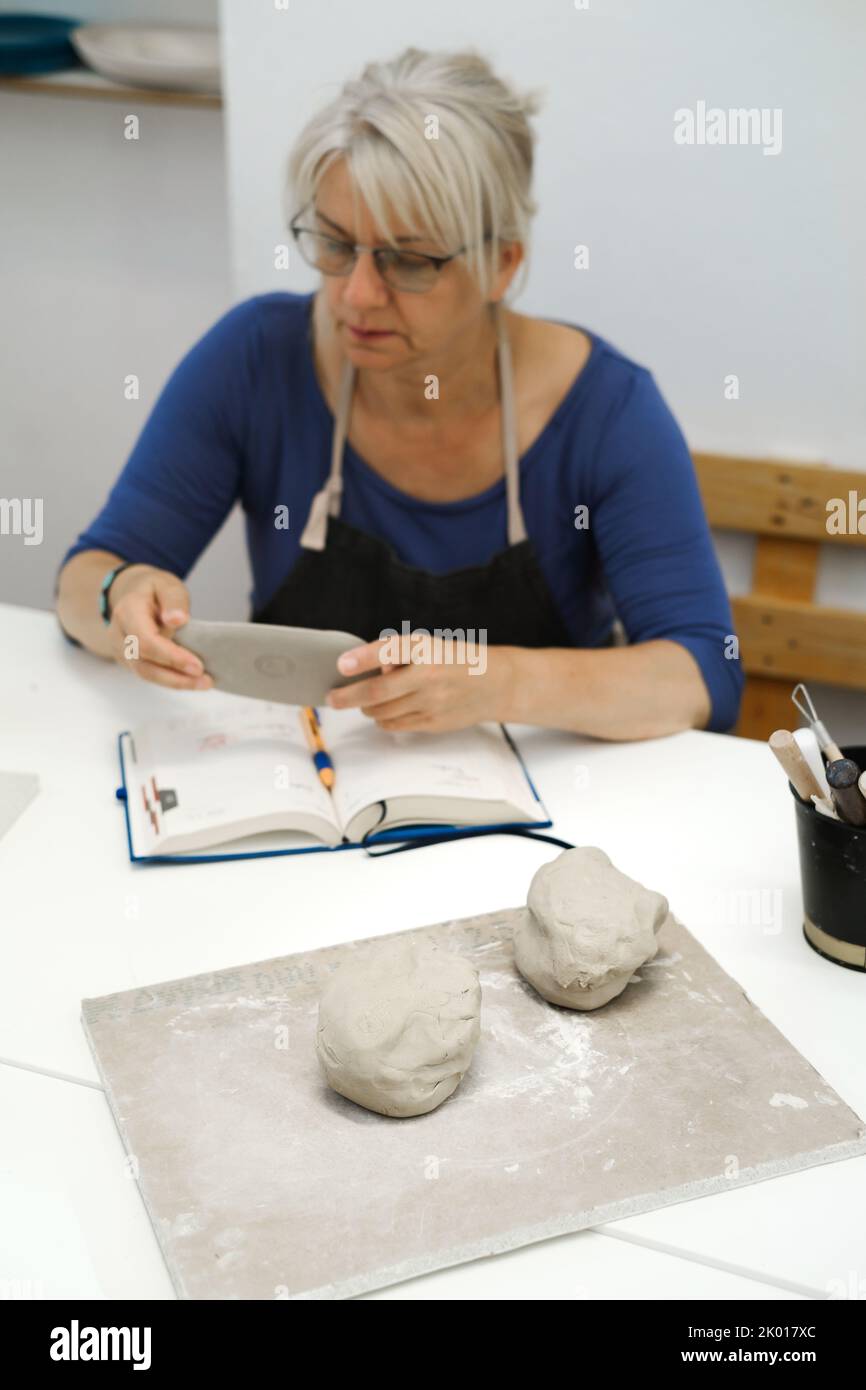 Frau Töpferin plant Arbeit in der Werkstatt, Schreiben in Notebook im Lichtstudio ihre Kreationen. Tonstücke auf dem Tisch. Kleines kreatives Unternehmen. Kopieren Stockfoto