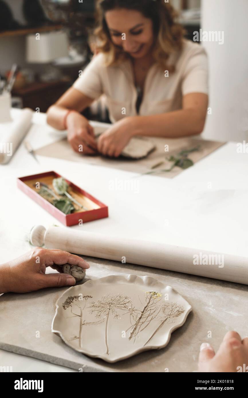 Frau, die mit Blumen gerollt Ton, so dass Keramikplatte im Studio mit Blumenmuster. Handgefertigte kreative Arbeiten. Töpferwerkstatt für Erwachsene. Stockfoto