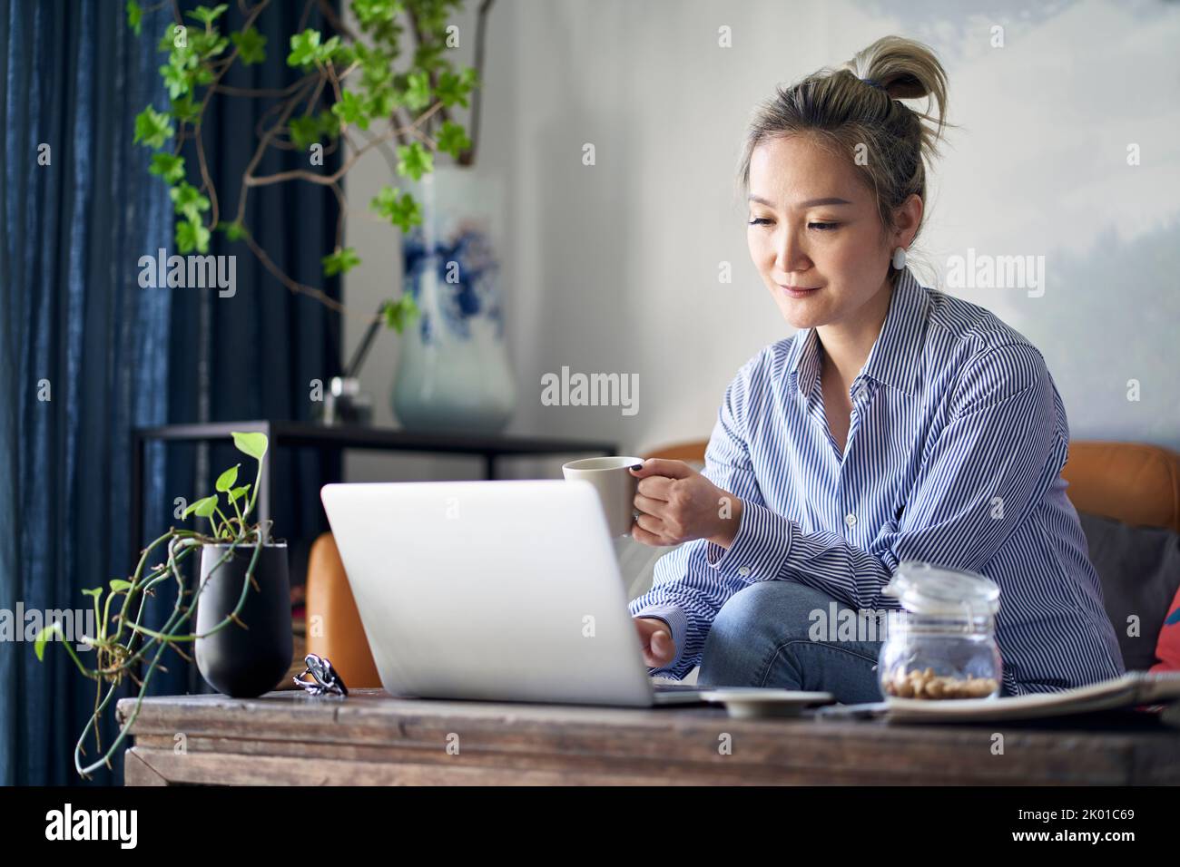 Reife professionelle asiatische Frau, die von zu Hause aus auf der Couch sitzt und Kaffee trinkt, während sie auf einen Laptop schaut Stockfoto