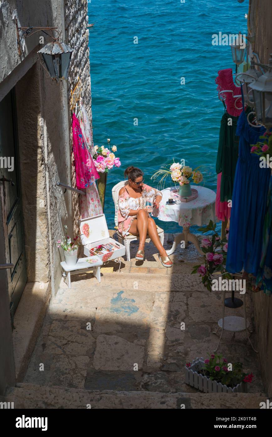Nett gekleidete Frau, die sich die Stufen einer Gasse in der Nähe des Wassers an einem Tisch mit bunten Blumen setzt und sich in Rovinj, Kroatien, kleidet Stockfoto