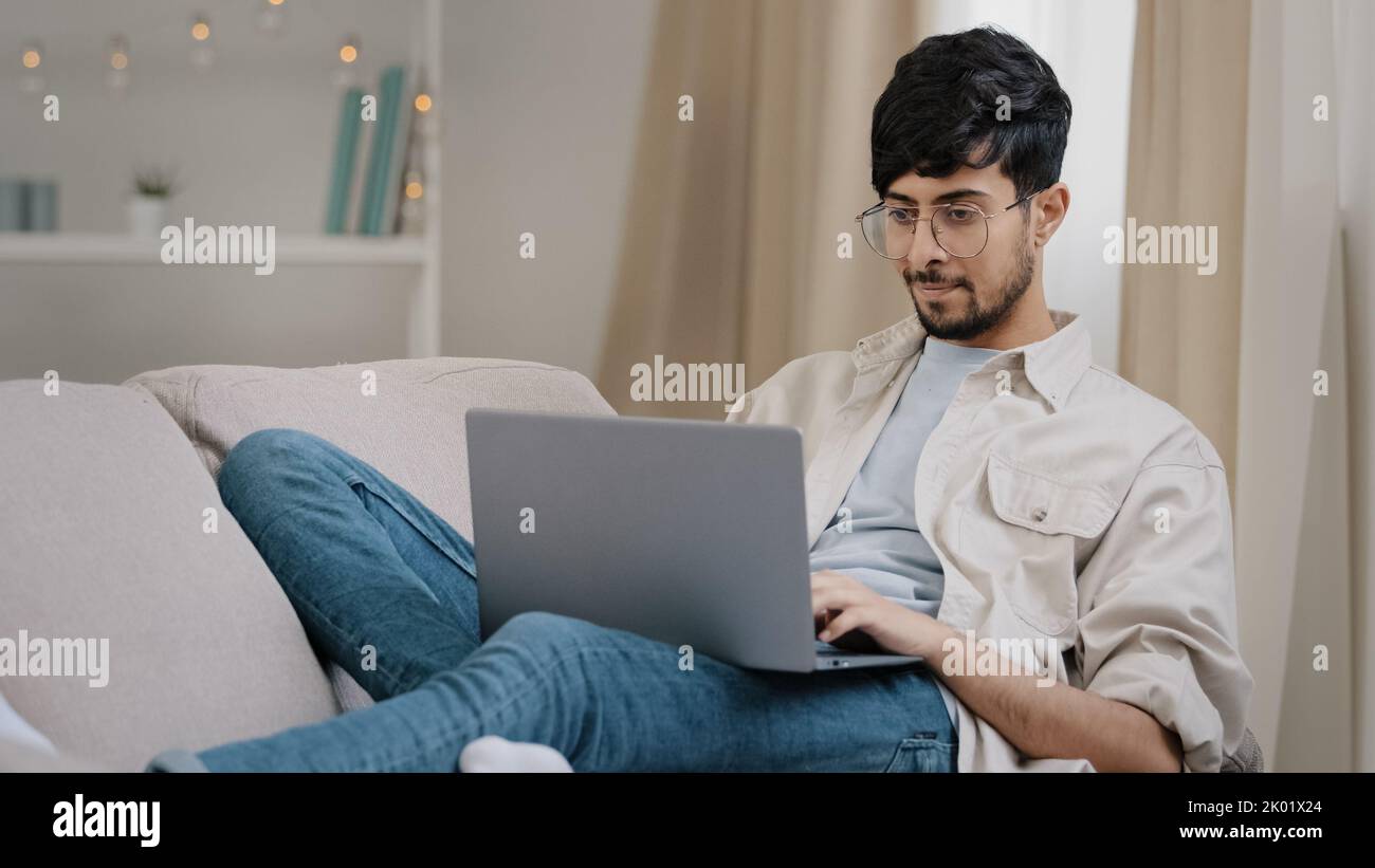 Junger Mann arab Freiberufler Geschäftsmann männlich bärtig Student in Brille liegt auf der Couch zu Hause mit Laptop arbeiten studieren online denken Stockfoto