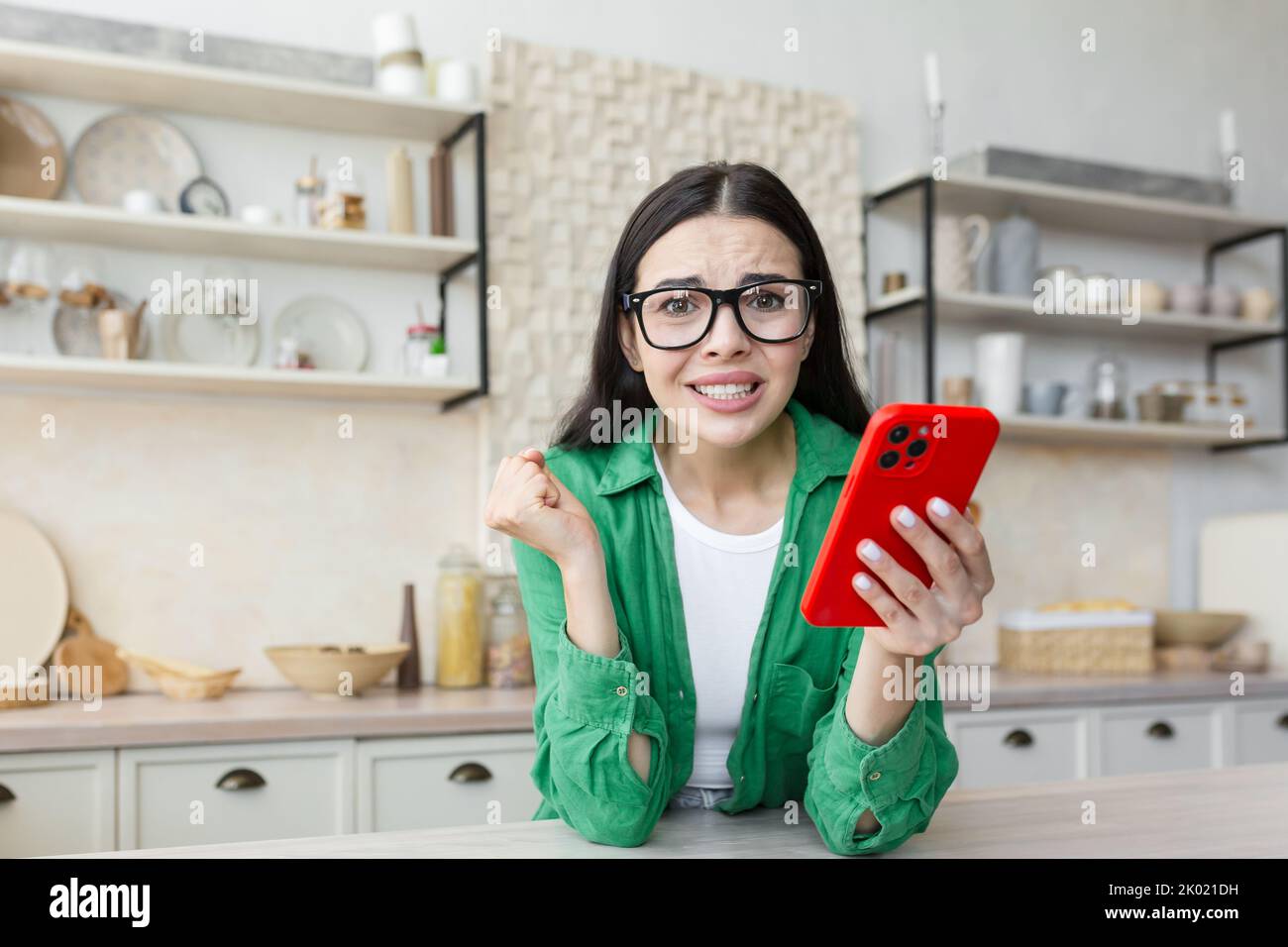 Nervöse junge Frau in einer Brille, die ein rotes Telefon hält und sich auf einen wichtigen Anruf oder eine wichtige Nachricht freut. Zu Hause in der Küche, auf die Kamera schauend, besorgt. Stockfoto