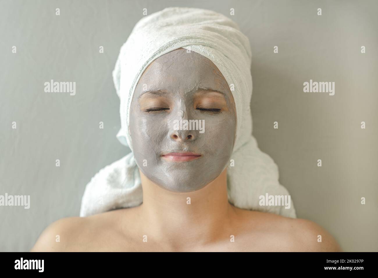 Junge Frau mit Tonmaske auf dem Gesicht und Handtuch auf dem Kopf in Form von Turban liegt mit geschlossenen Augen und genießt eine kosmetische Spa-Behandlung für die Gesichtshautpflege. Nahaufnahme des Gesichts, Draufsicht. Stockfoto