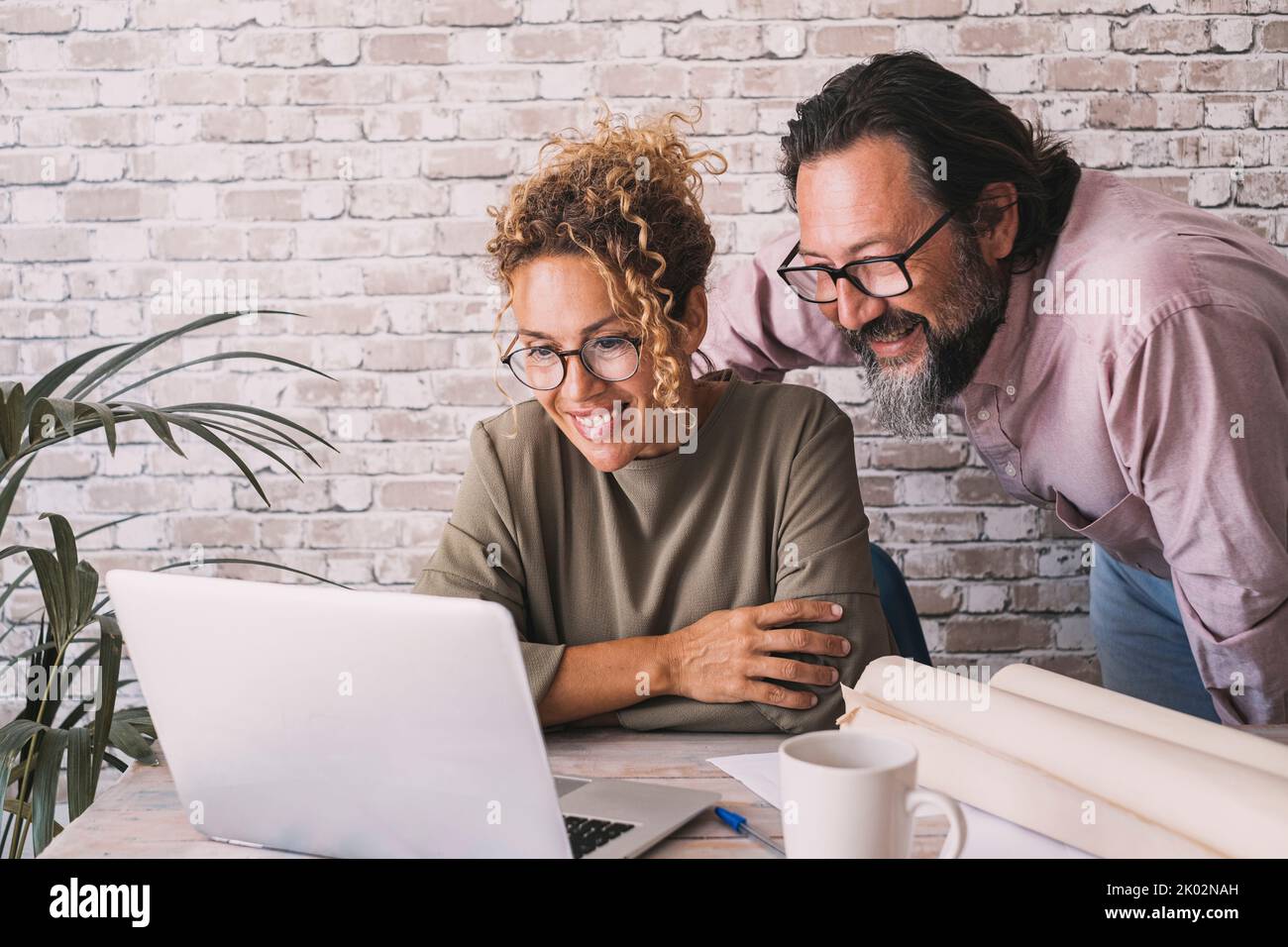 Mann und Frau arbeiten im Büro mit Laptop- und Papierprojekten zusammen. Moderne Geschäftswelt und E-Commerce-Job-Lifestyle. Glückliches Paar, das auf einem Computerbildschirm aussuckt und lächelt. Konzept eines Online-Jobanrufs Stockfoto