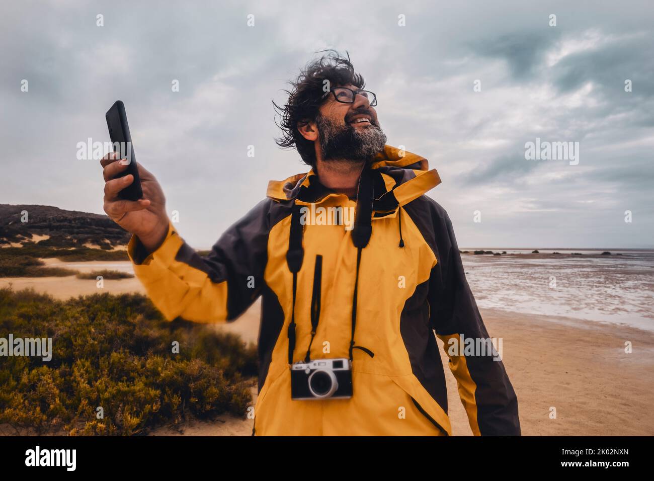 Porträt eines wilden Fotografen, der schlechtes Wetter am Strand genießt. Fröhlicher Tourist mit Fotokamera und Handy. Strand und Meer und Wolken im Hintergrund. Freizeitaktivitäten im Freien in der Natur Stockfoto
