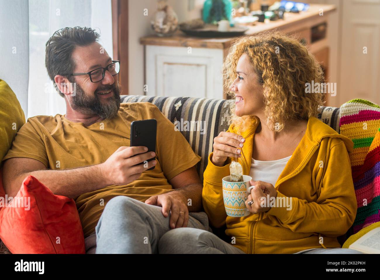 Erwachsene Paare genießen Freizeit zu Hause beim Frühstück am Morgen. Mann am Telefon und Frau, die Tee trinkt. Menschen zusammen in Beziehung und im echten häuslichen Leben sitzen auf dem Sofa in der Wohnung Stockfoto