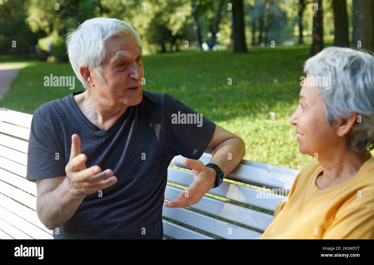 Paare mittleren Alters, multiethnische Ehepartner sitzen auf der Bank Genießen Sie Gespräche im öffentlichen Sommerpark, während Sie freundliche Gespräche führen und dabei Gedanken oder Nachrichten teilen Stockfoto