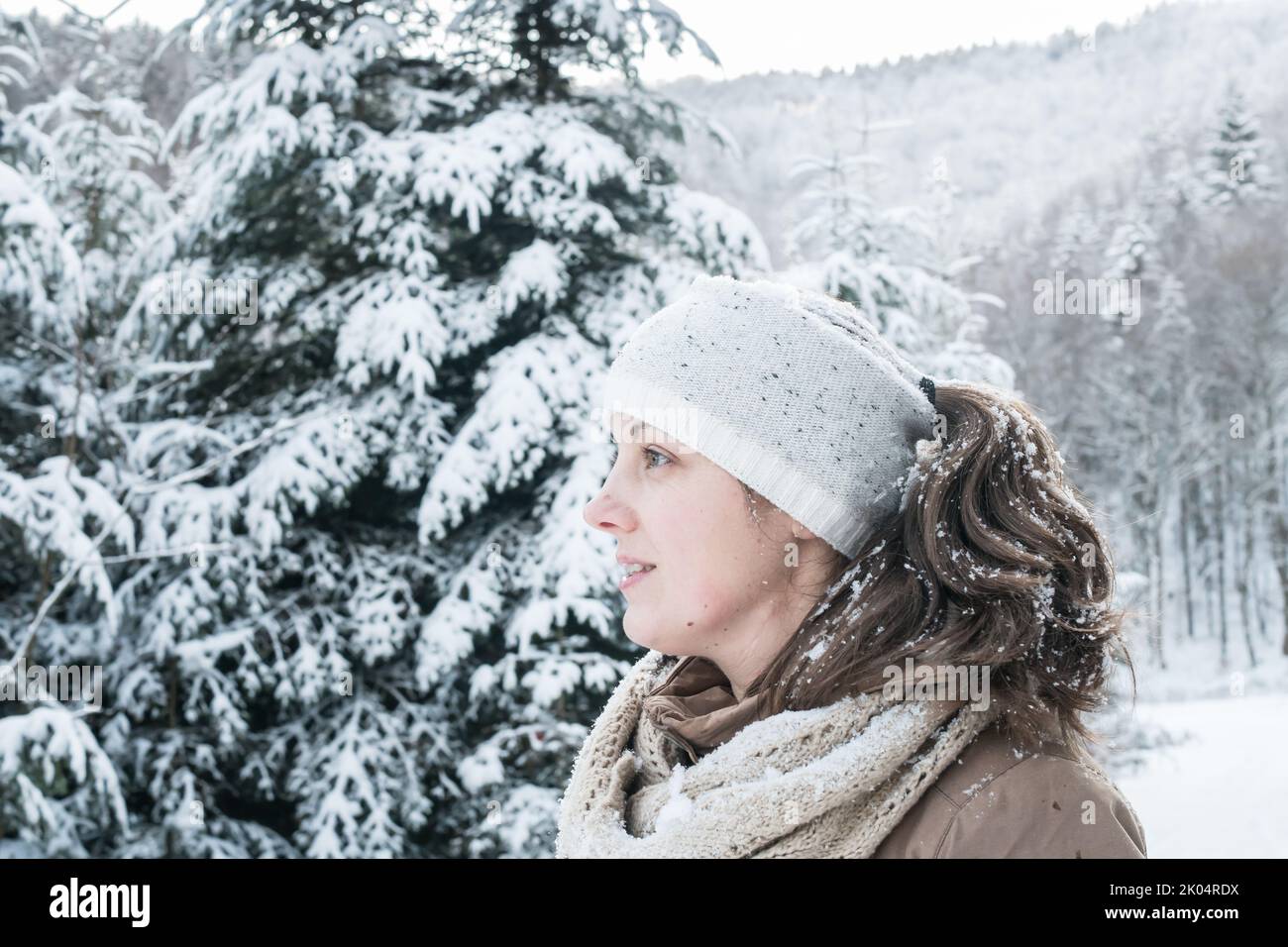 Lächelnde 30-jährige Frau, die auf den schneebedeckten Winterwald blickt. Sie trägt Stirnband, Schal und Handschuhe. Glückliche 30 Jahre alte kaukasische Dame. Stockfoto