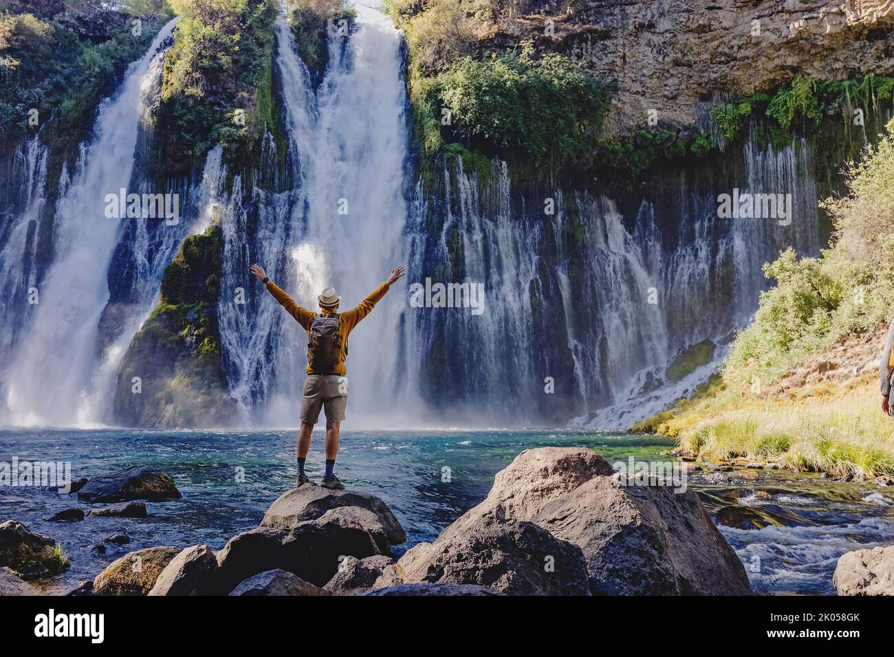 Tourist steht mit erhobenen Händen mit Blick auf den schönen Wasserfall, Blick von hinten Stockfoto