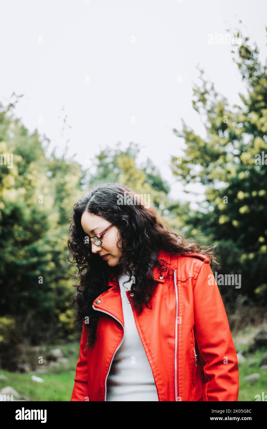 Die lockige, brünette lateinerin blickt nach unten und trägt in der Natur eine rote Lederjacke. Stockfoto