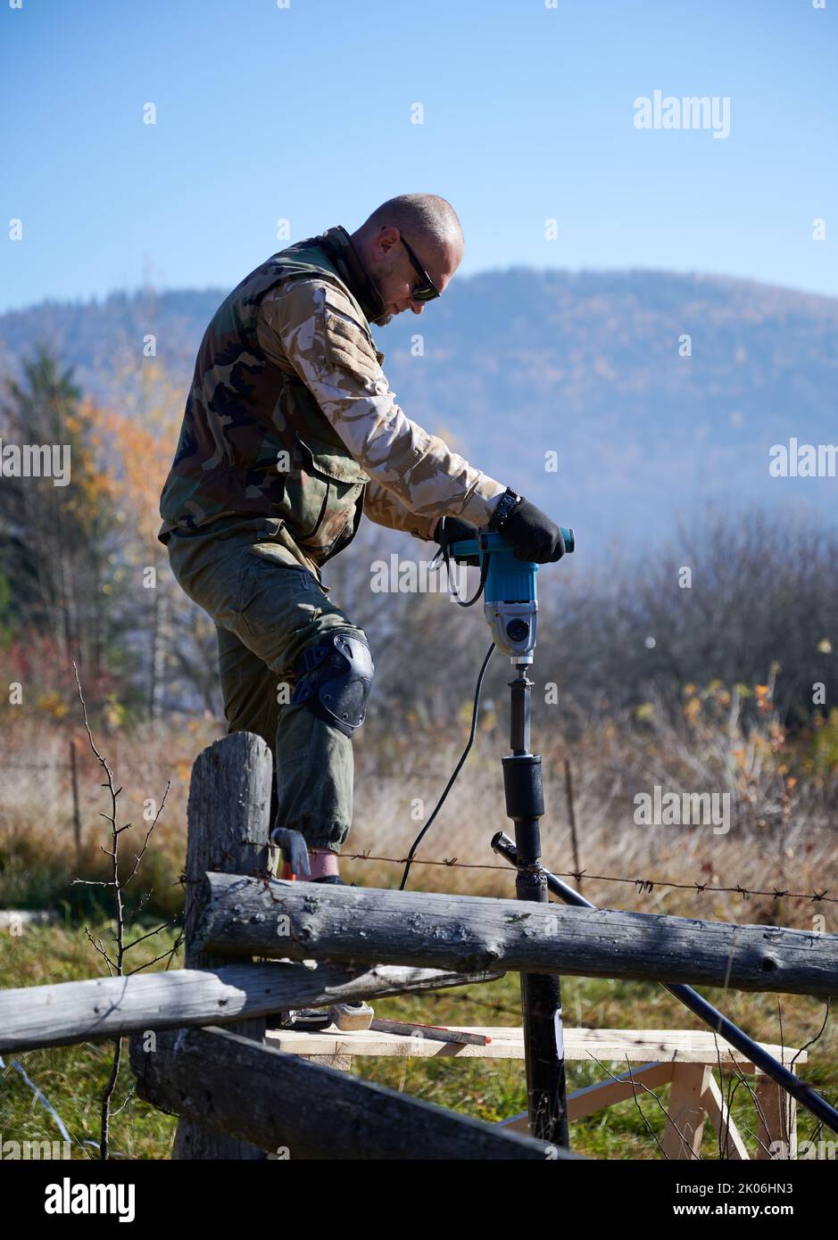 Männlicher Arbeiter Gebäude Pfahlfundament für Holzrahmenhaus. Der Baumeister bohrt Stapel auf blauem Himmel in den Boden. Stockfoto