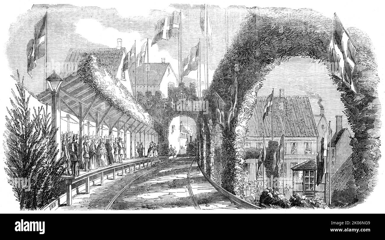 Bahnhof Flensburg, 1854. Eröffnung der in Großbritannien gebauten Königlich Dänischen Eisenbahn durch Friedrich VII., König von Dänemark. Bei der Eröffnung waren Samuel Morton Peto, ein Partner der Firma Peto, Brassey und Betts, und der Ingenieur George Parker Bidder anwesend. "...seine Majestät... empfing die englische Eisenbahngesellschaft mit großer Höflichkeit, umgeben von seinem Stab; und nachdem er zum Ausdruck gebracht hatte, wie sehr er von der unerwarteten Schnelligkeit, mit der die Eisenbahn fertiggestellt worden war, zufrieden war, ging er zum Bahnhof...". Die Plattform, die hier mit viel Grün und einem Triumphbogen mit den Initialen des Königs zu sehen ist, Stockfoto