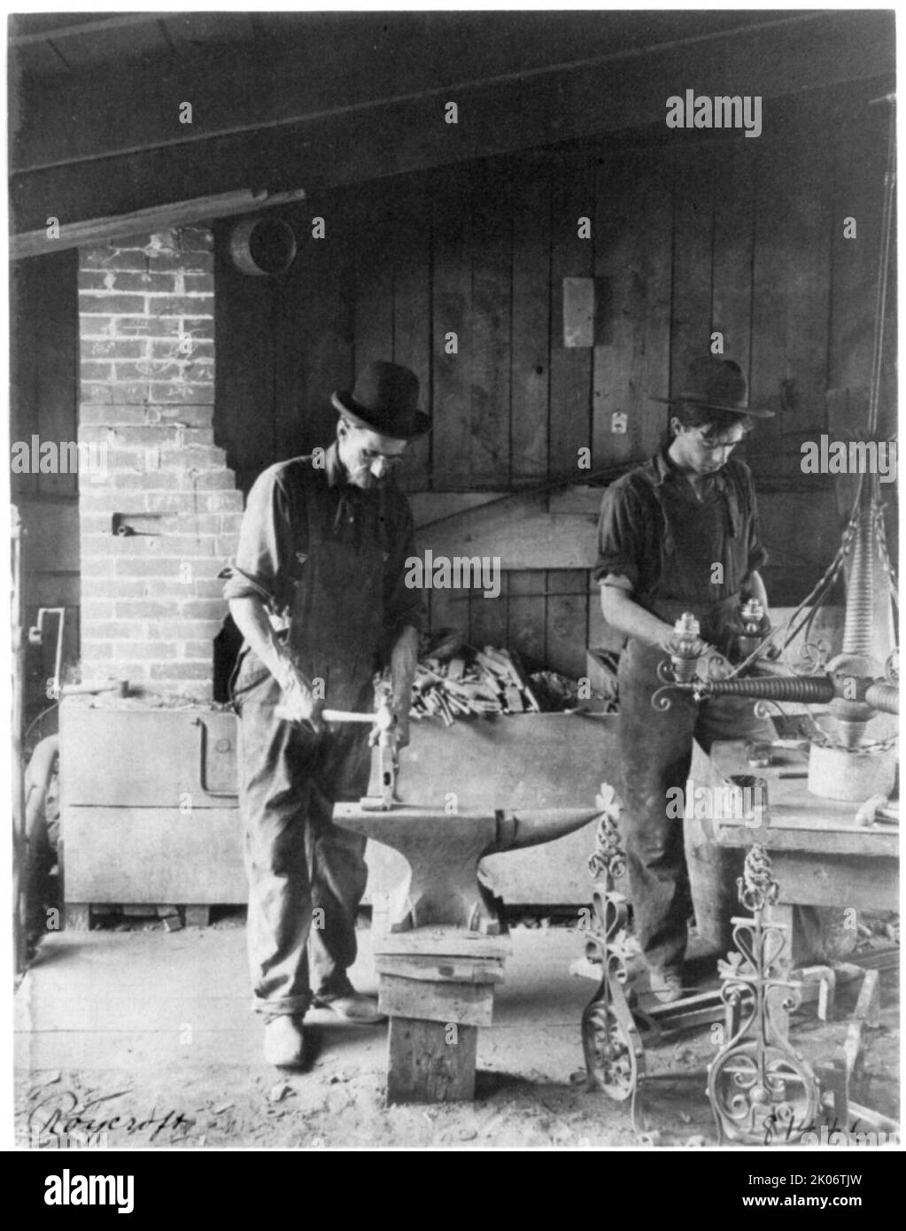 Roycroft Shop, East Aurora, N.Y., 1900. 2 Männer arbeiten am Amboss und am Tisch mit schmiedeeisernem Kronleuchter in der Schmiede Roycroft (erbaut 1899), Grove Street, East Aurora, New York. Stockfoto