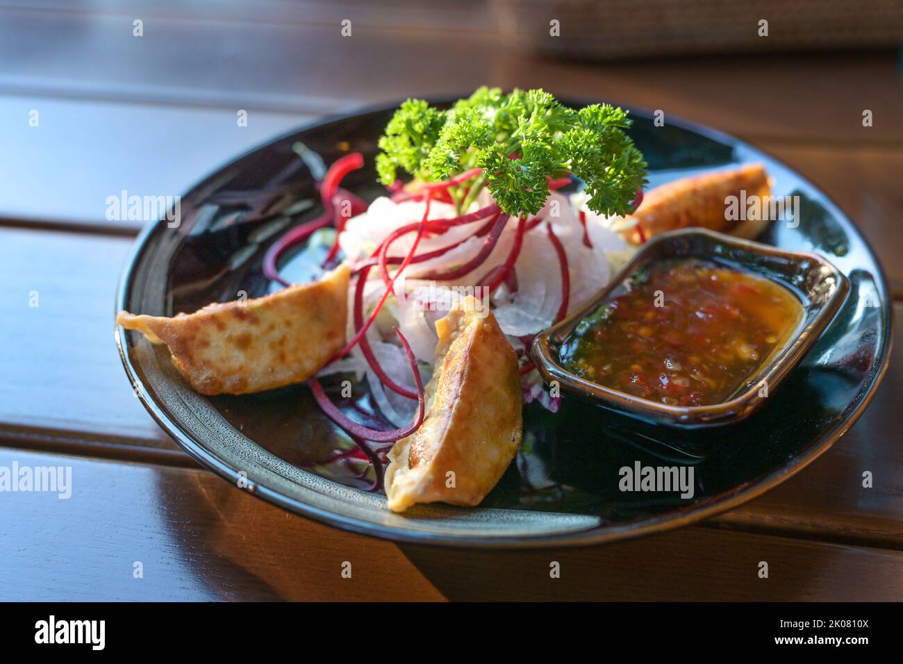 Frittierte Gyoza serviert mit würziger süß-saurer Sauce und Salat als Vorspeise Gericht auf einem schwarzen Teller, asiatische Speisen auf einem Holztisch, Platz kopieren, auswählen Stockfoto