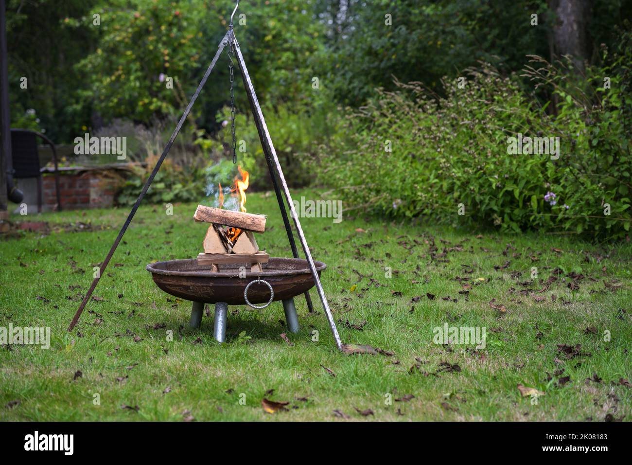 Gestapeltes Brennholz in einer Feuerschale brennt mit Flammen und raucht unter einem Dreibeingrill in einem Garten, Vorbereitung für eine Grillparty, Kopierplatz, ausgewählt Stockfoto