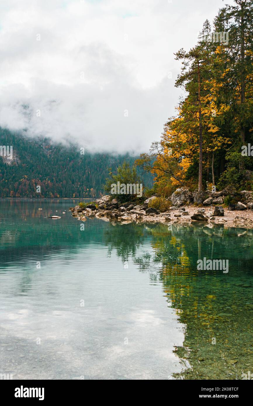 Gemütlicher Herbstmorgen in den bayerischen Bergen, Deutschland. Alpenlandschaft mit Seenküste, kleinem Dorf, Wolken, Wald und Reflexion im Wasser Stockfoto