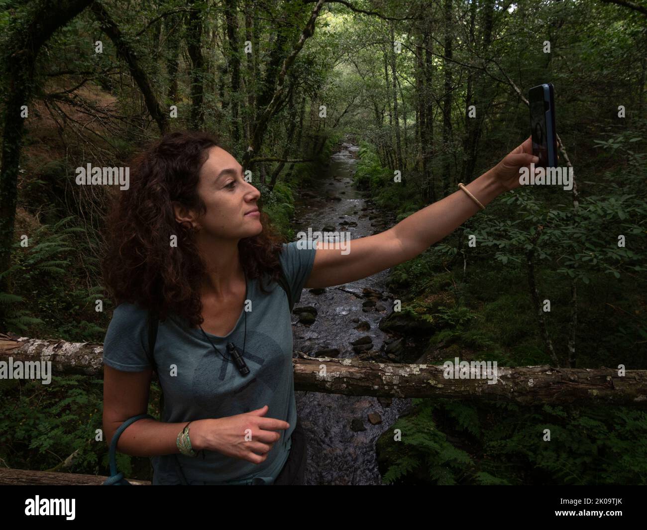 Brünette Frau, die ein Selfie auf einer Holzbrücke mit Fluss fließt durch eine Eiche und Kastanien im Hintergrund. Stockfoto