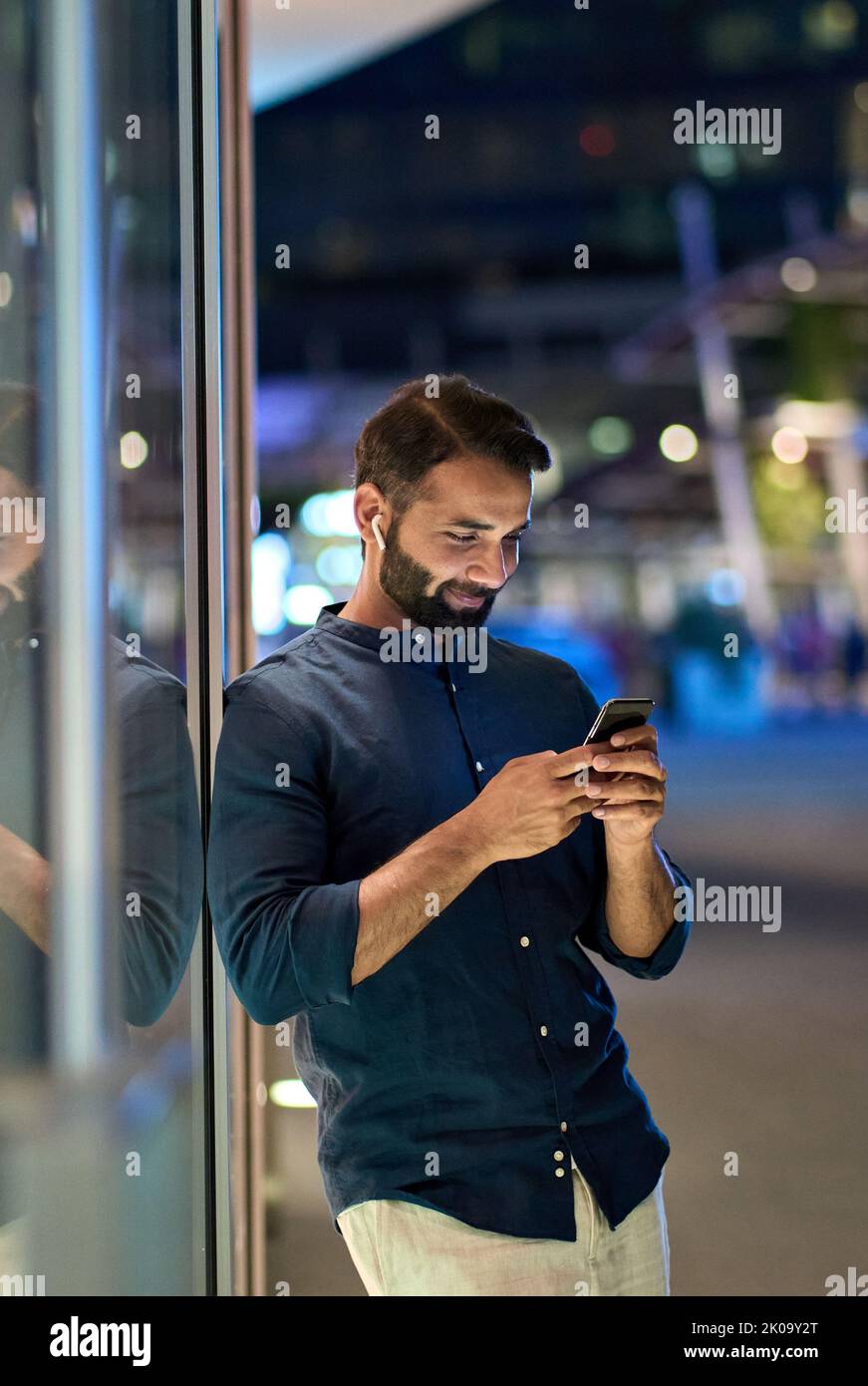 Indischer Mann mit Ohrstöpsel, der nachts sein Mobiltelefon im Freien hält, vertikal. Stockfoto