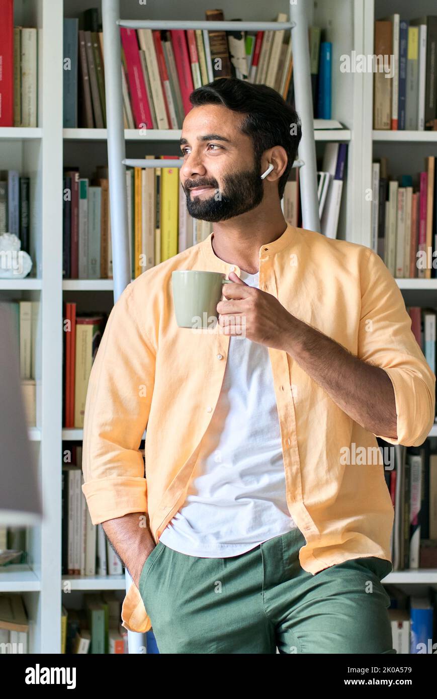 Nachdenklicher indischer Mann, der in Ohrstöpseln steht und Kaffee trinkt und Musik hört. Stockfoto