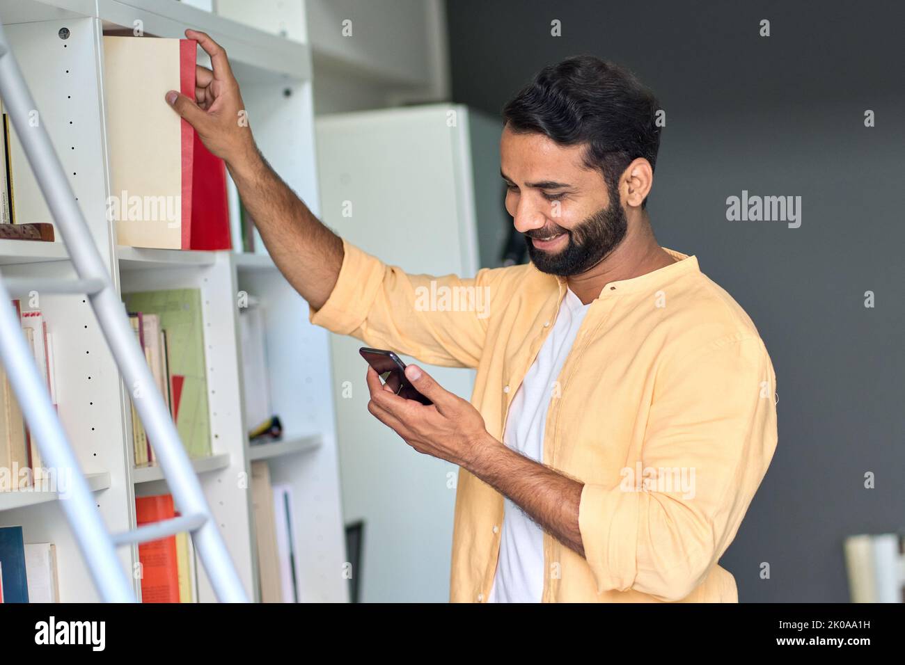 Ein indischer Mann, der ein Telefon benutzt, steht zu Hause und nimmt ein Buch aus den Bücherregalen. Stockfoto