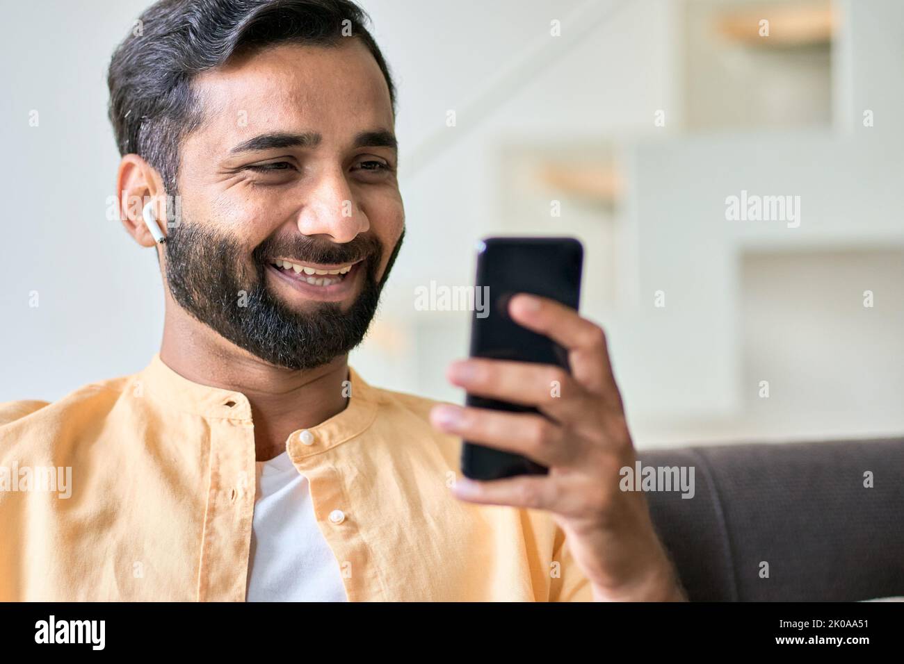 Glücklicher indischer Mann, der zu Hause mit einem Ohrstöpsel auf dem Smartphone sitzt. Stockfoto