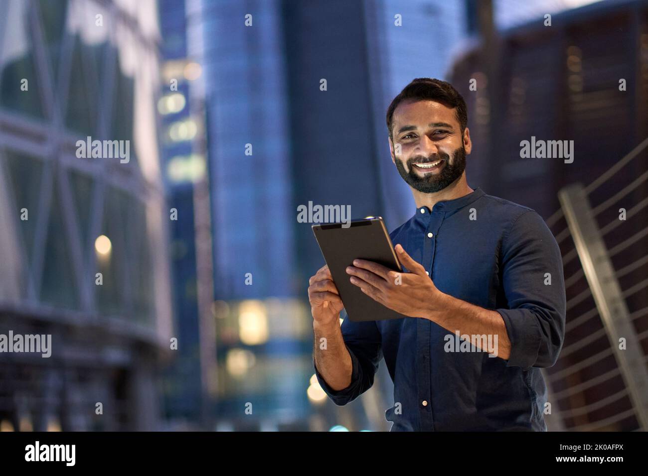 Lächelnder indischer Geschäftsmann, der in der Nachtstadt mit einem digitalen Tablet steht. Stockfoto