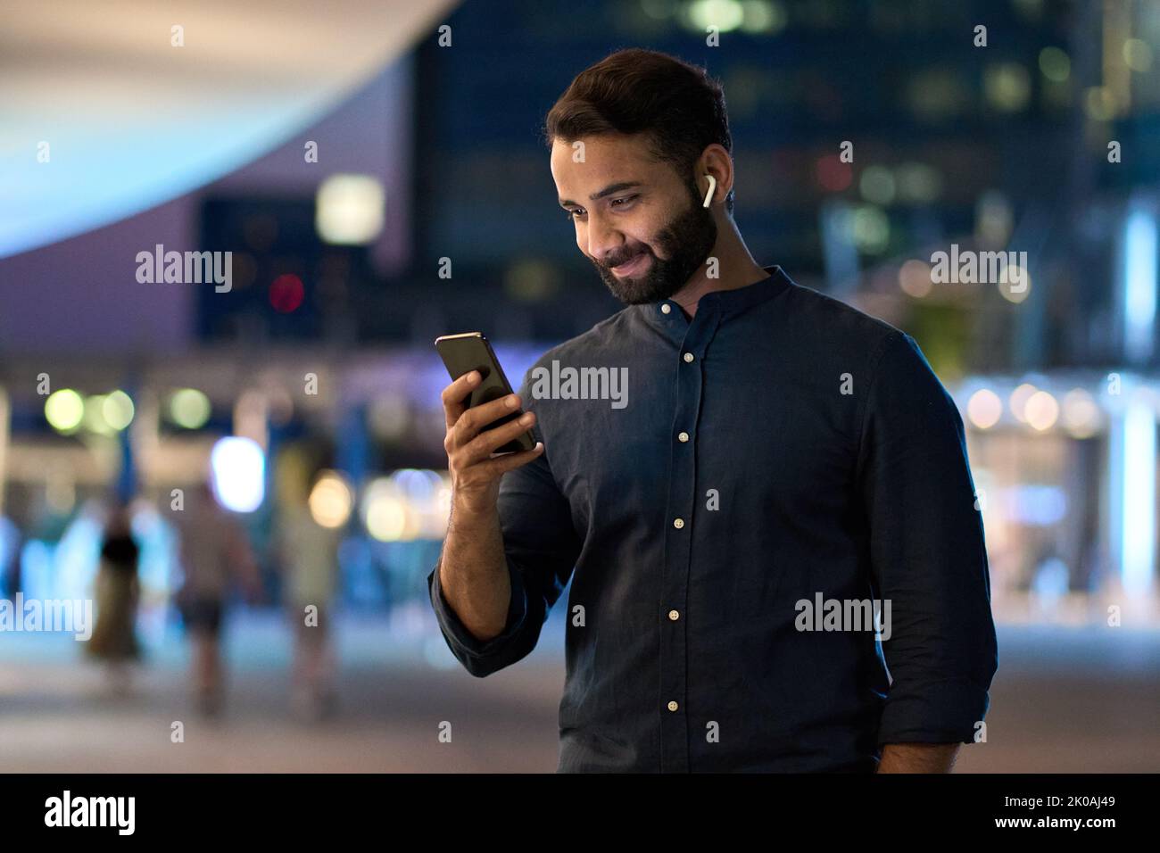 Indischer Geschäftsmann mit Ohrstöpsel, der in der Nachtstadt mit einem Smartphone steht. Stockfoto