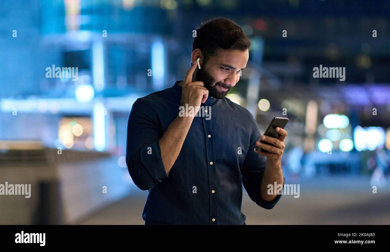 Indischer Mann mit Ohrstöpsel, der nachts mit dem Smartphone ein Handy hält. Stockfoto