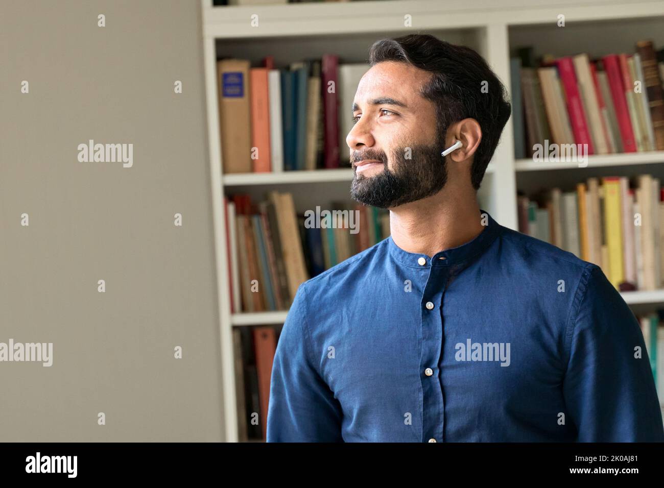 Nachdenklicher indischer Mann mit Ohrstöpseln, der Podcast hört und durch das Fenster schaut. Stockfoto