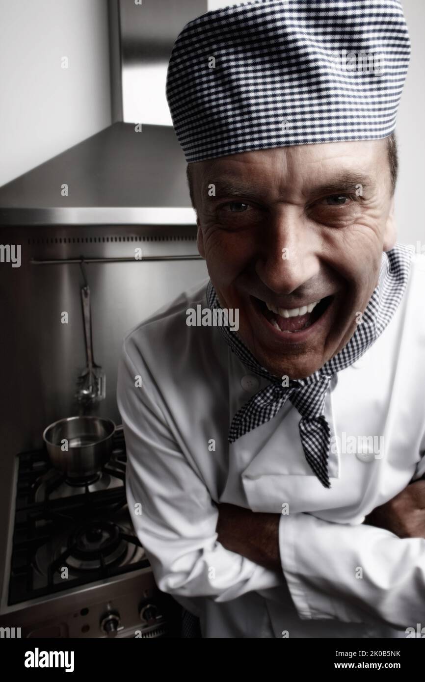 Lustig reifer Koch vor dem Gasofen und der Dunstabzugshaube stehen. Nahaufnahme eines lustigen, reifen Küchenchefs, der vor dem Gasherd und der Dunstabzugshaube steht. Stockfoto