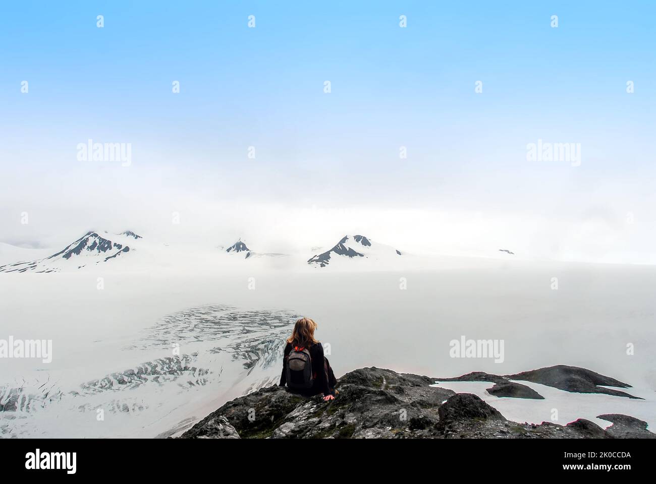 Eine Wanderin, die auf einem Berggipfel sitzt und über das Harding Ice Field in Alaska blickt und dabei die unglaublichen Aussichten und surrealen Aussichten genießt Stockfoto