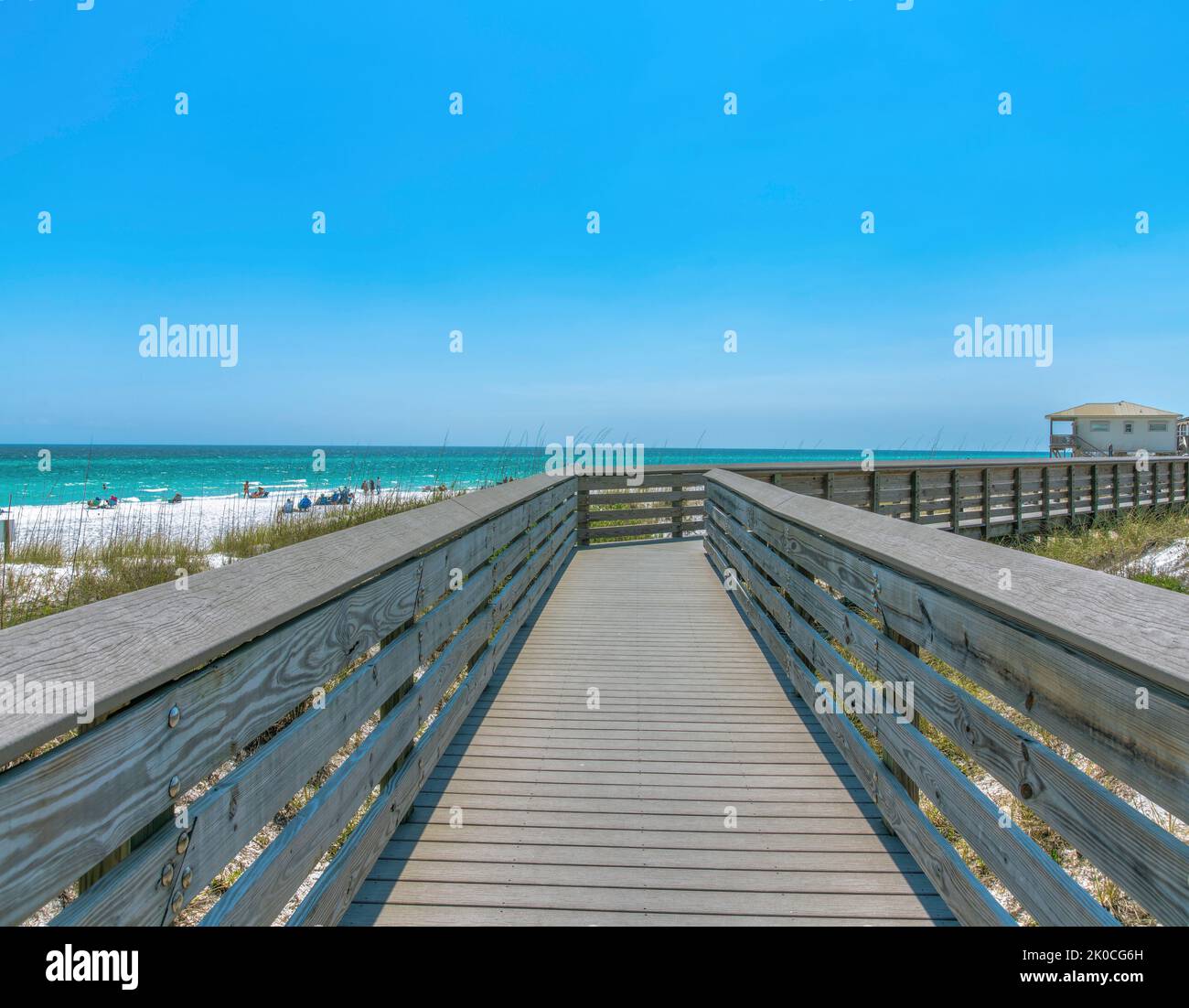 Hölzerne Promenade an einem Strand mit Menschen in Destin, Florida. Promenade mit Blick auf Gräser auf der Seite und Blick auf das blaue Wasser und Strandhaus Stockfoto