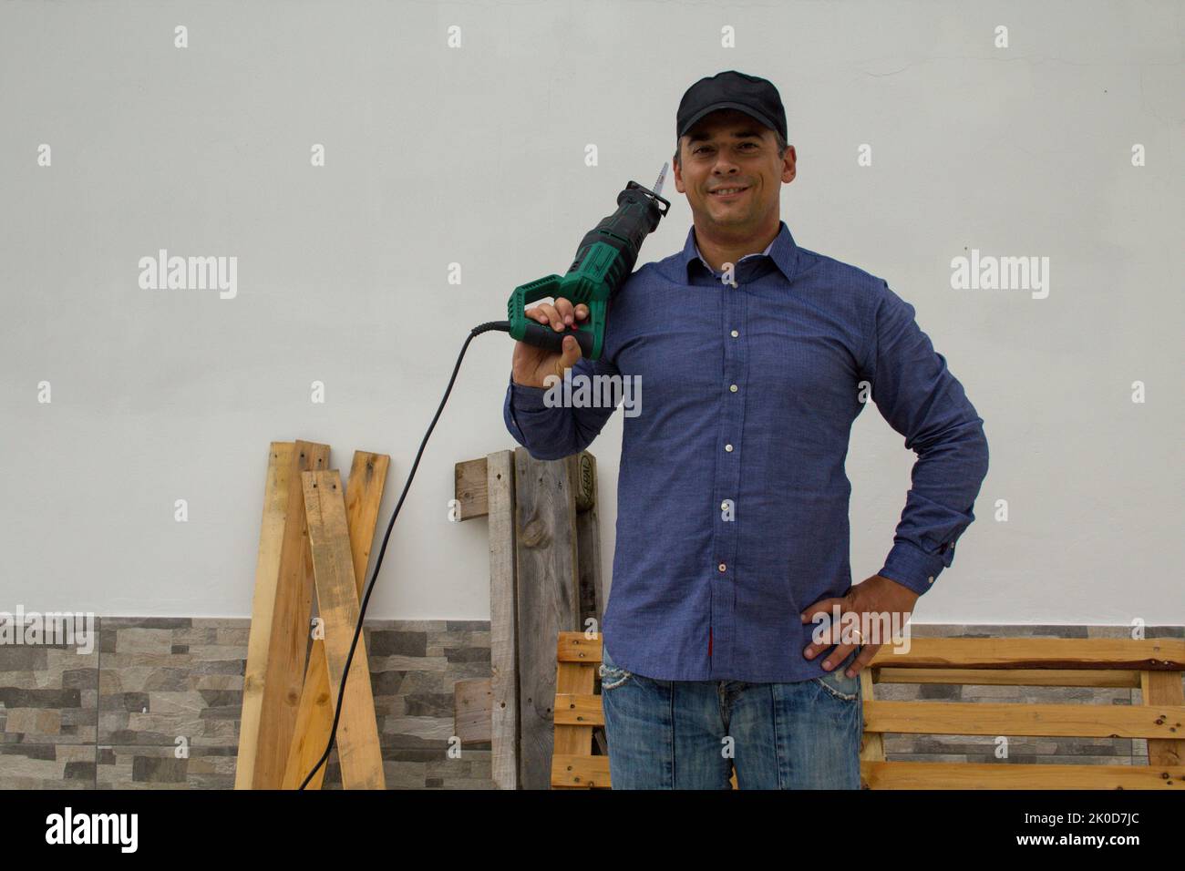 Lächelnder Handwerker mit einer Säge auf der Schulter beim Schneiden und Demontieren von Holzpaletten und Plattformen. Wiederverwendung von Holz für Do-it-yourself-Arbeiten Stockfoto