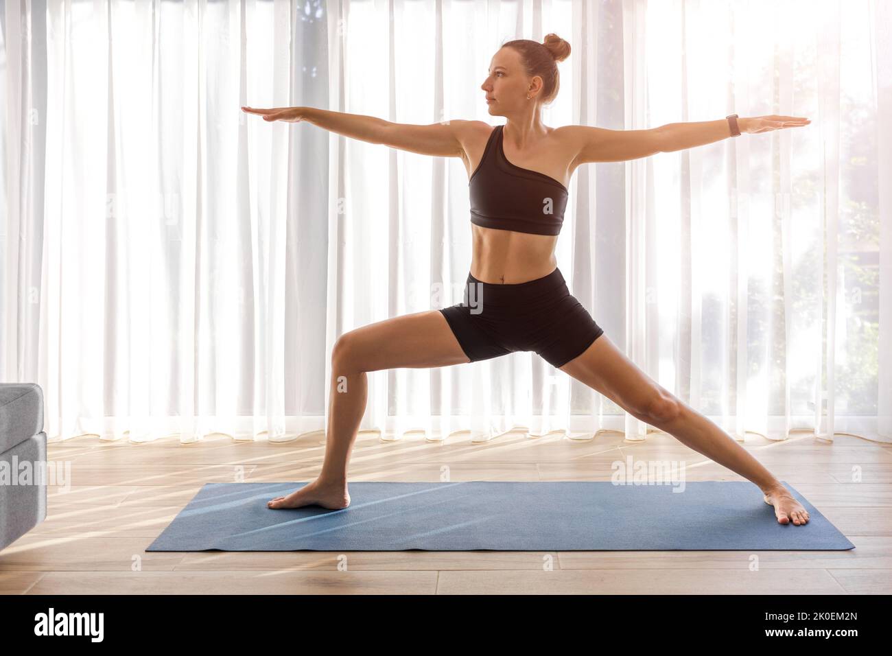 Junge Dame in schwarzer Sportkleidung, die im Zimmer mit großem Fenster Yoga macht Stockfoto